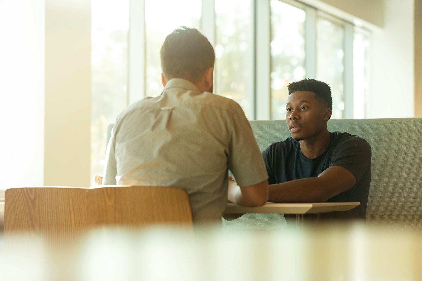 Two people in conversation at a table by a window, one listening while the other speaks